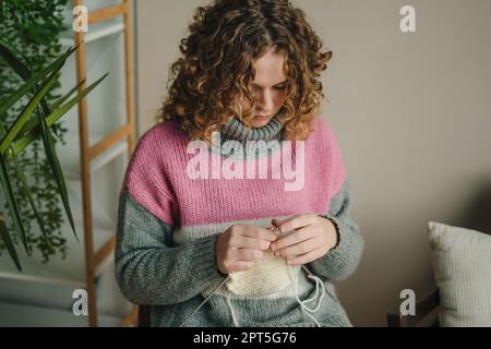 Calm curly-haired woman knitting with white thread sitting on sofa in cozy wooden interior house. Crafting and handmade. Lifestyle and beautiful Stock Photo