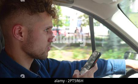 Young handsome serious man driving a car and typing on smartphone while looking at road. Side view. Close up of male driver using mobile phone while Stock Photo