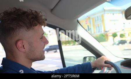 Close up shot of happy Caucasian man driving a car and smiling. Cheerful positive guy with curly hair drives his auto looking at road. Side view Stock Photo