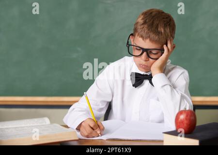 Not another test. A young boy with glasses and a bow-tie sitting in class writing despondently in a book Stock Photo