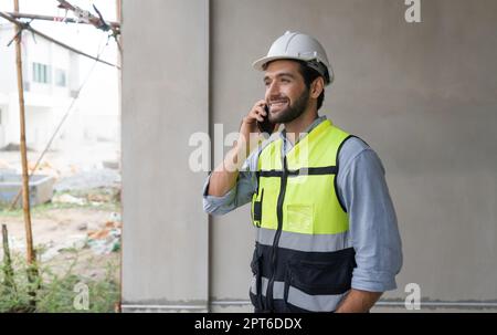 Young engineer in hardhat and safety vest talking on mobile phone. Work environment of engineers at the construction site. Stock Photo
