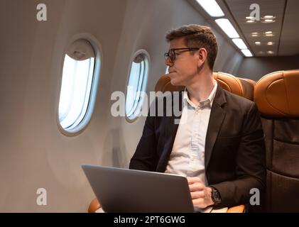 Businessman in formal wear and glasses looking out of aircraft window during flight  while working with laptop computer. Internet connection on board Stock Photo
