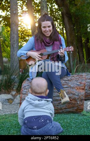 Woman playing ukulele smiling and a baby sitting on the grass watching her as a spectator Stock Photo