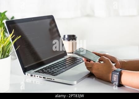 Programmer woman looking mobile phone program code at workplace, software developer hands typing data coding programming javascript with laptop comput Stock Photo