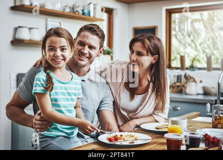 She truly is the apple of their eye. Portrait of a little girl having breakfast with her parents at home. Stock Photo