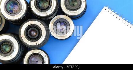 Several photographic lenses and white notebook lie on a bright blue background. Space for text Stock Photo