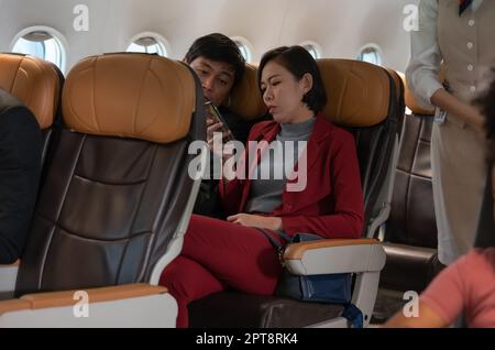 Couple passengers sitting on seat and using mobile phone in airplane before flight. Woman holding and showing somethings on smartphone to her boyfrien Stock Photo
