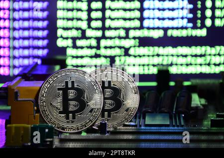 Two bitcoins lies on a videocard surface with background of screen display of cryptocurrency mining by using the GPUs Stock Photo