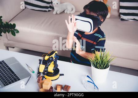 Virtual Reality Simulator. Asian kid boy using VR glasses on robotic arm in workshop, Child learning programer control robot arm with sensors to pick Stock Photo