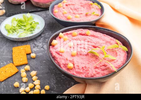 Hummus with beet and microgreen basil sprouts in ceramic bowl on a black concrete background and orange textile. Side view, close up. Stock Photo