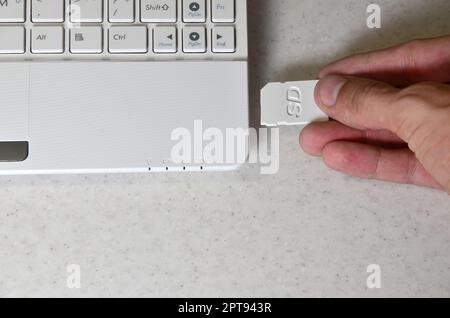 A male hand inserts a white compact SD card into the corresponding input in the side of the white netbook. Man uses modern technologies to store memor Stock Photo