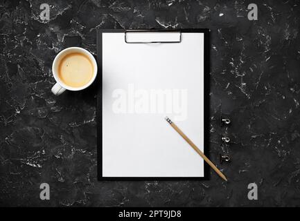 Photo of clipboard with blank letterhead, coffee cup, pencil and clips on black plaster background. Blank stationery set. Flat lay. Stock Photo