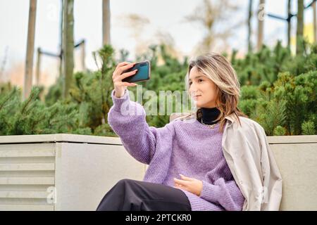 Cute young woman makes selfie or video call while sitting on bench in the city Stock Photo