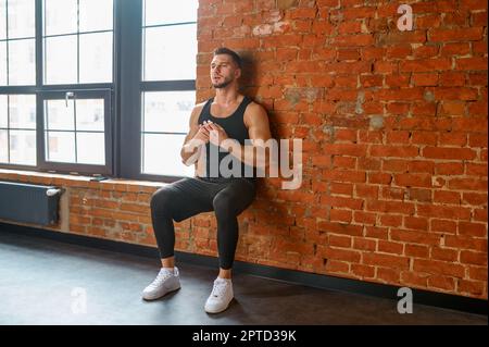 Athlete doing training exercise against gym wall. Sportsman exercising squats leaning back on brickwall. Sport, bodybuilding, fitness and people conce Stock Photo