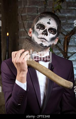 Man in suit with sugar skull makeup playing with axe. Face painting art. Stock Photo