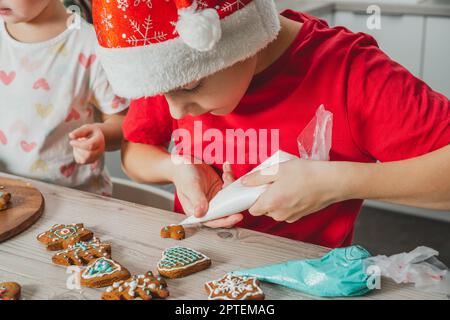 Boy 8 years old in red Santa hat decorate Christmas gingerbread cookies with icing on wooden table in kitchen. Merry Christmas, Happy New Year Stock Photo