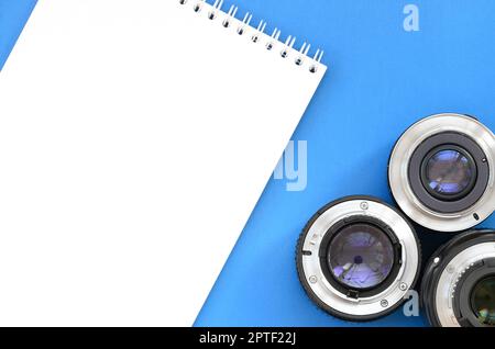 Several photographic lenses and white notebook lie on a bright blue background. Space for text Stock Photo