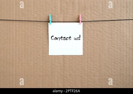 Contact us! Note is written on a white sticker that hangs with a clothespin on a rope on a background of brown cardboard Stock Photo