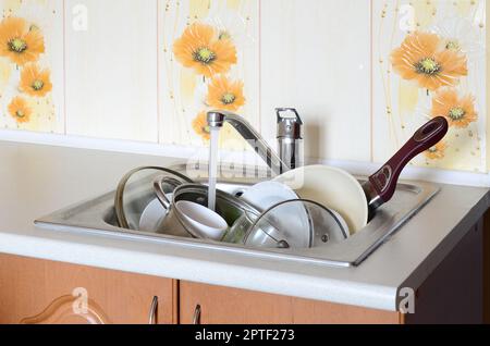 Dirty dishes and unwashed kitchen appliances lie in foam water under a tap from a kitchen faucet Stock Photo