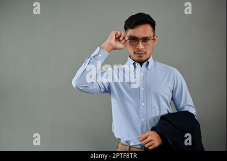 Professional millennial Asian businessman or male boss touching his eyeglasses, holding his suit, and standing on an isolated grey background. Stock Photo