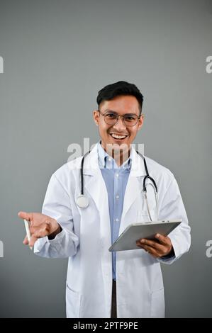Smart and handsome millennial Asian male doctor in a uniform holding a digital tablet while standing on an isolated grey background. presenting, givin Stock Photo