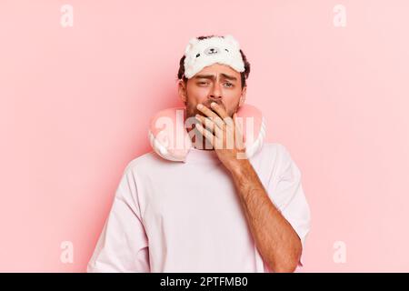 Funny sleepy man in white T-shirt with sleep mask on his head just woke up, stands against pink background, yawns and covers his mouth with hand, slee Stock Photo