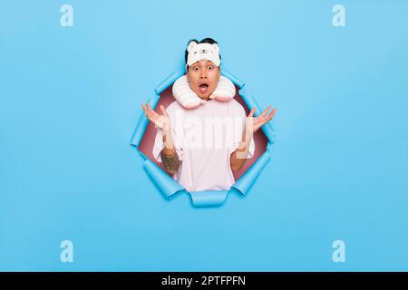 Portrait of excited young man in white T-shirt wearing sleep mask on his forhead and inflatable sleeping pillow around his neck, he poses in the middl Stock Photo