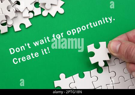 Don't wait for opportunity. Create it. The hand folds a white jigsaw puzzle and a pile of uncombed puzzle pieces lies against the background of the gr Stock Photo