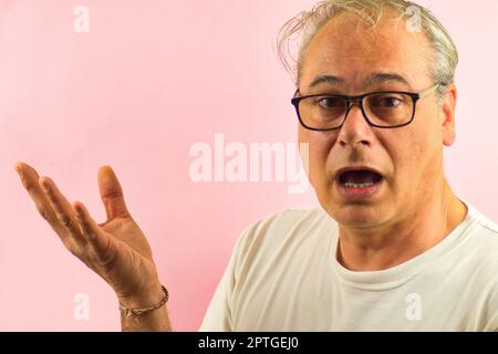 mature man with glasses and grey hair shows surprised something with outstretched hand on pink background Stock Photo