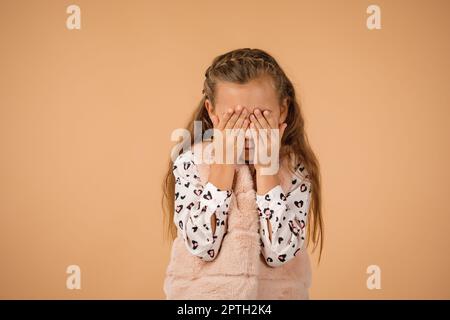 Cute little child girl closes her eyes with her hand on beige background. I see nothing Stock Photo