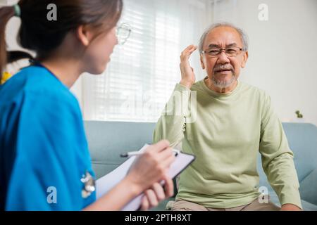 Asian elderly man patient have headache and female nurse talking, checking up and recording current symptoms on clipboard in living room, Home healthc Stock Photo