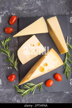 Various types of cheese with rosemary and tomatoes on black slate board on a black concrete background. Top view, flat lay, close up. Stock Photo