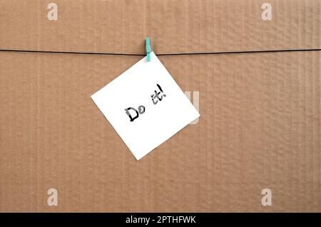 Do it! Note is written on a white sticker that hangs with a clothespin on a rope on a background of brown cardboard Stock Photo
