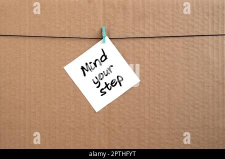 Mind your step. Note is written on a white sticker that hangs with a clothespin on a rope on a background of brown cardboard Stock Photo