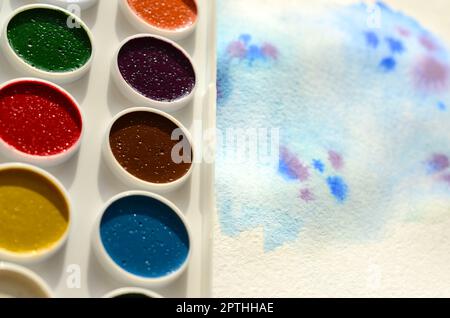 A new set of watercolors lies on a sheet of paper, which shows an abstract watercolor drawing in the form of blue strokes. The concept of amateur pain Stock Photo