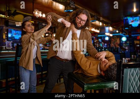 Young woman trying to separate craze mad fighting friends. Conflict of football fans at sport bar Stock Photo