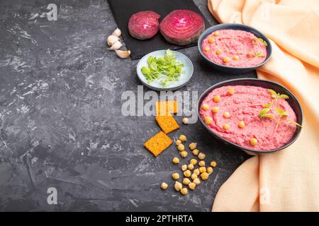 Hummus with beet and microgreen basil sprouts in ceramic bowl on a black concrete background and orange textile. Side view, copy space. Stock Photo