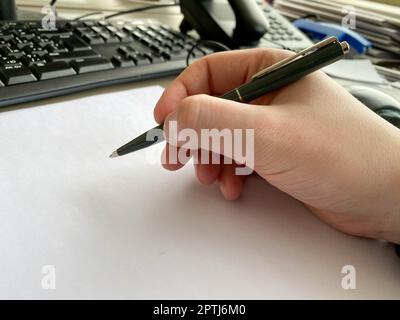 A man's hand in a shirt and with a fitness bracelet holds a pen and writes on the table at the office table with a computer with a keyboard. Business Stock Photo