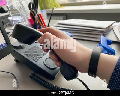 A man's hand in a shirt holds a landline phone receiver on a work desk with office supplies in a business office. Stock Photo