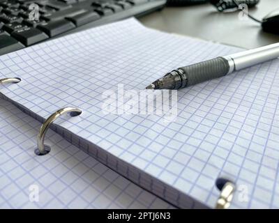 A writing pen rests on a notepad with squared paper sheets on a work desk with stationery in a business office. Stock Photo