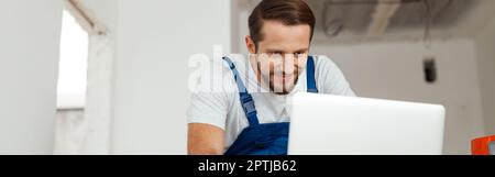 Professional handyman in workwear leaning on hands and smiling while using laptop, standing indoors Stock Photo