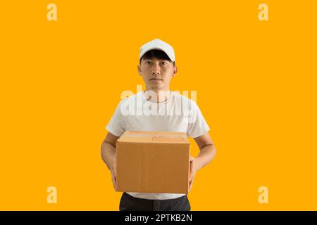 Young Asian delivery man wearing cap and white blank t-shirt holding parcel post box isolated on yellow background. express delivery service concept. Stock Photo