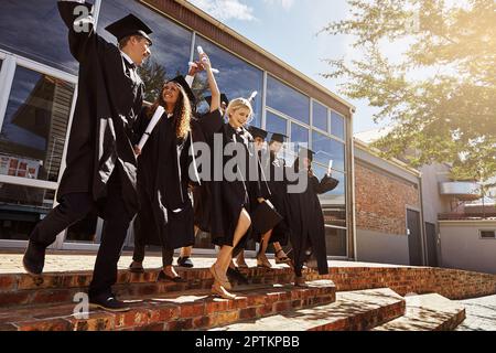 Celebrating their achievements together. a group of smiling university students jumping in the air and cheering outside on graduation day. Stock Photo