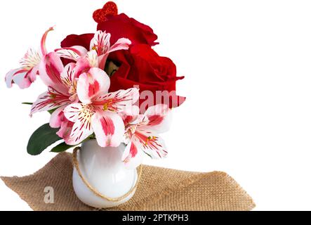 red roses and pink flowers in a small bouquet and a part of a red glitter heart can be seen, and a small white circular vase on a jute fabric. Stock Photo
