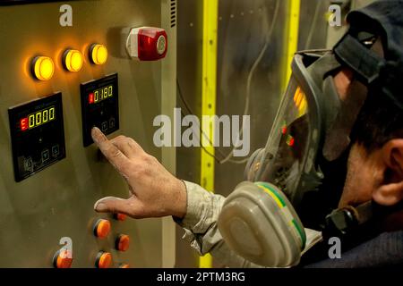 a white man in a work mask stands at the machine and presses the buttons to turn on the furnace for painting metal, work is being done in the workshop Stock Photo
