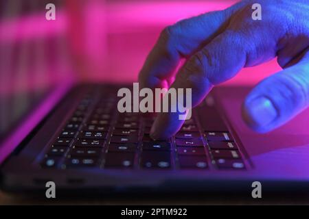 Person typing at laptop computer at night. Male uses keyboard, shakes buttons with fingers. Internet security, cyber attack. Trendy neon light. Creati Stock Photo