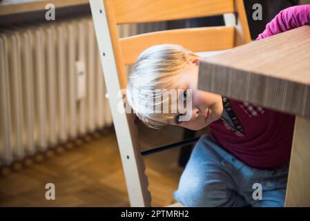 Little boy curiously looking from under the edge of the table, Munich, Germany Stock Photo