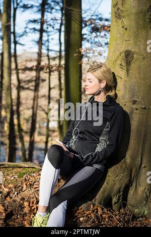 Young female jogger having a break while listening music on smart phone in forest Stock Photo