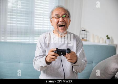 Asian senior old man enjoying holding joystick playing video game at home in living room, mature man hands using game controller, Funny on retirement Stock Photo