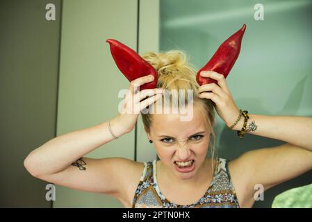 Portrait of a beautiful young woman making horns with two paprika on her head, Munich, Bavaria, Germany Stock Photo
